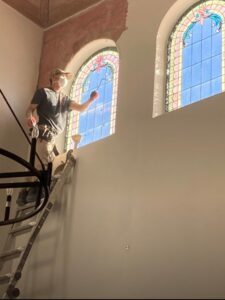 Man working on a tall ladder near arched windows in a sunlit room.